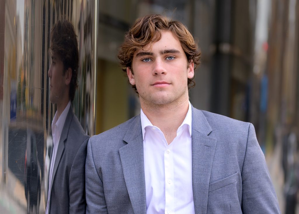 High school senior young man in a gray suit and white shirt stands confidently against a shiny building surface, reflecting his image. Urban setting, calm expression.