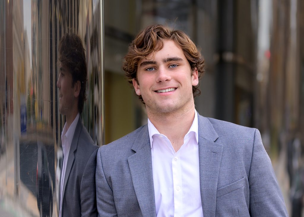 High school senior young man in a gray suit and white shirt stands smiling against a shiny building surface, reflecting his image. Urban setting, calm expression.