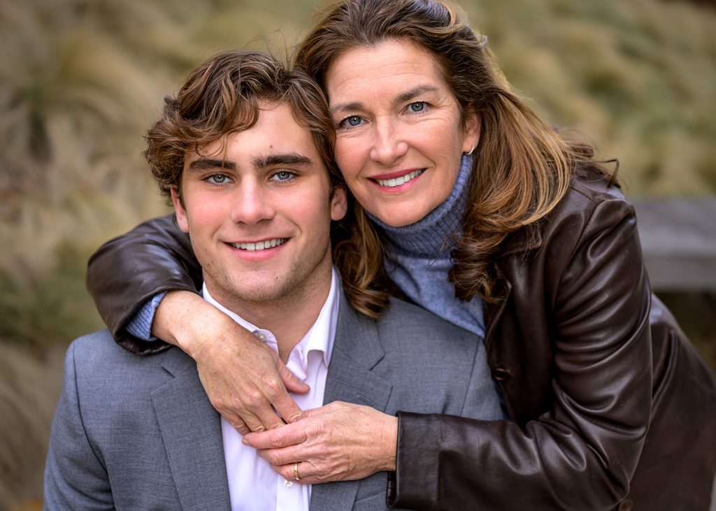 Mom hugging her son from behind, during his senior photo session with J. Hill Photography. They are outdoors, wearing a brown jacket and grey suit, conveying a sense of closeness and happiness.