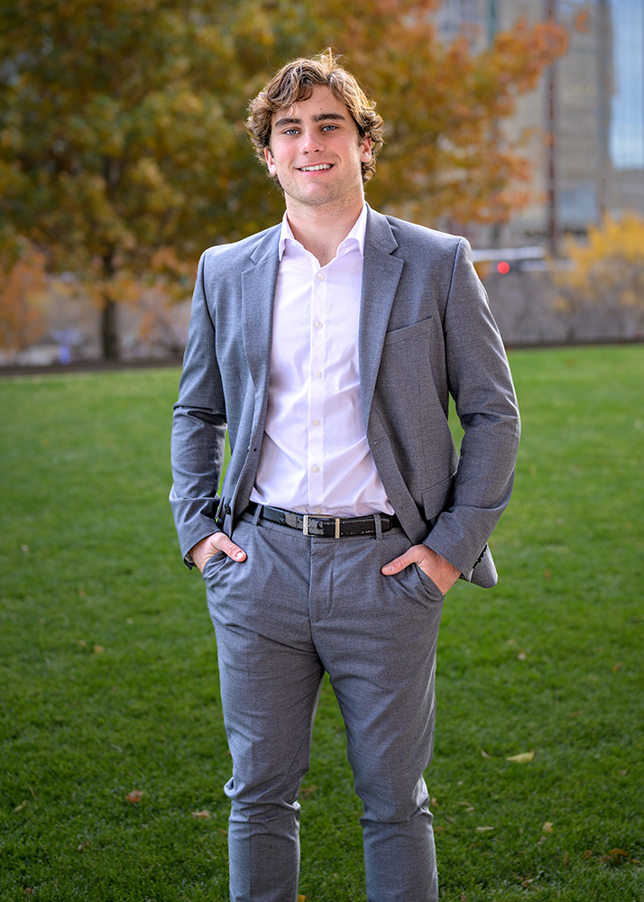 A high school senior boy in a gray suit and white shirt stands confidently on green grass, hands in pockets. Autumn trees and a building are blurred in the background.