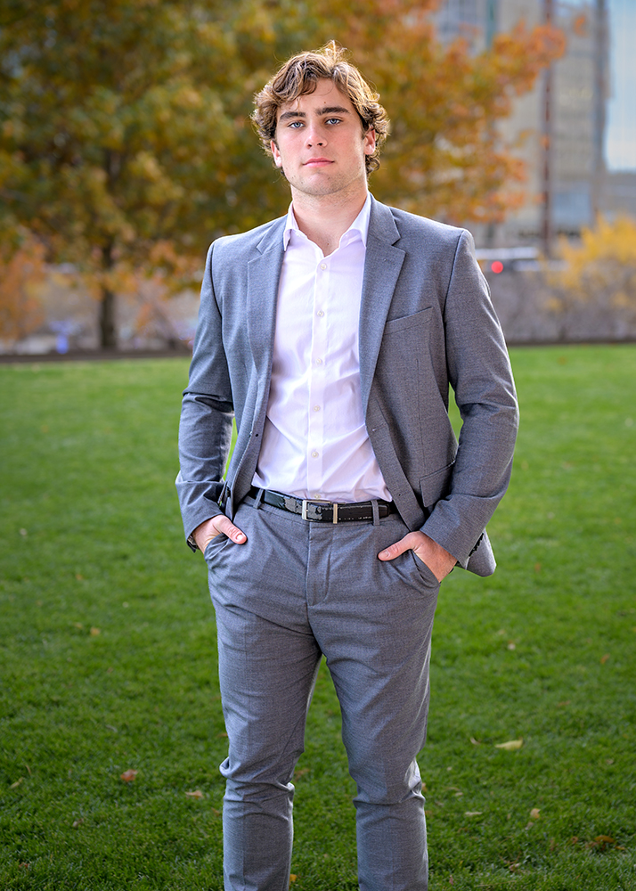 A senior guy in a gray suit and white shirt stands confidently on green grass, hands in pockets. Autumn trees and a building are blurred in the background.