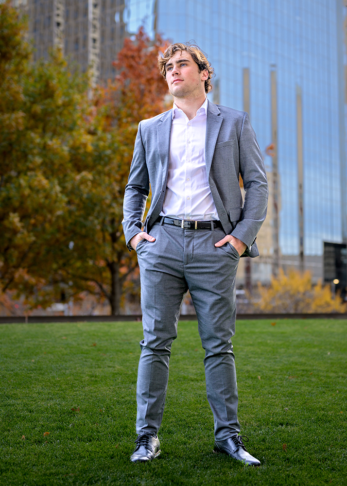 A confident high school senior in a gray suit and white shirt stands on a green lawn. Autumn trees and a tall glass building create an urban backdrop.