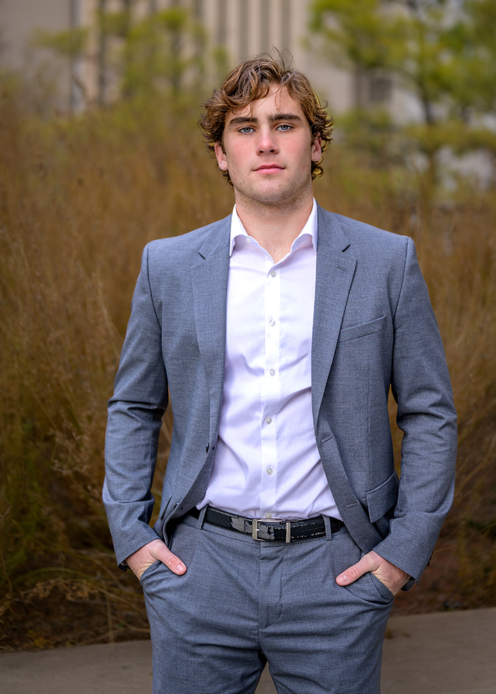 Senior boy in a gray suit and white shirt stands outdoors, hands in pockets. Background features soft-focus greenery and a neutral building, conveying confidence.