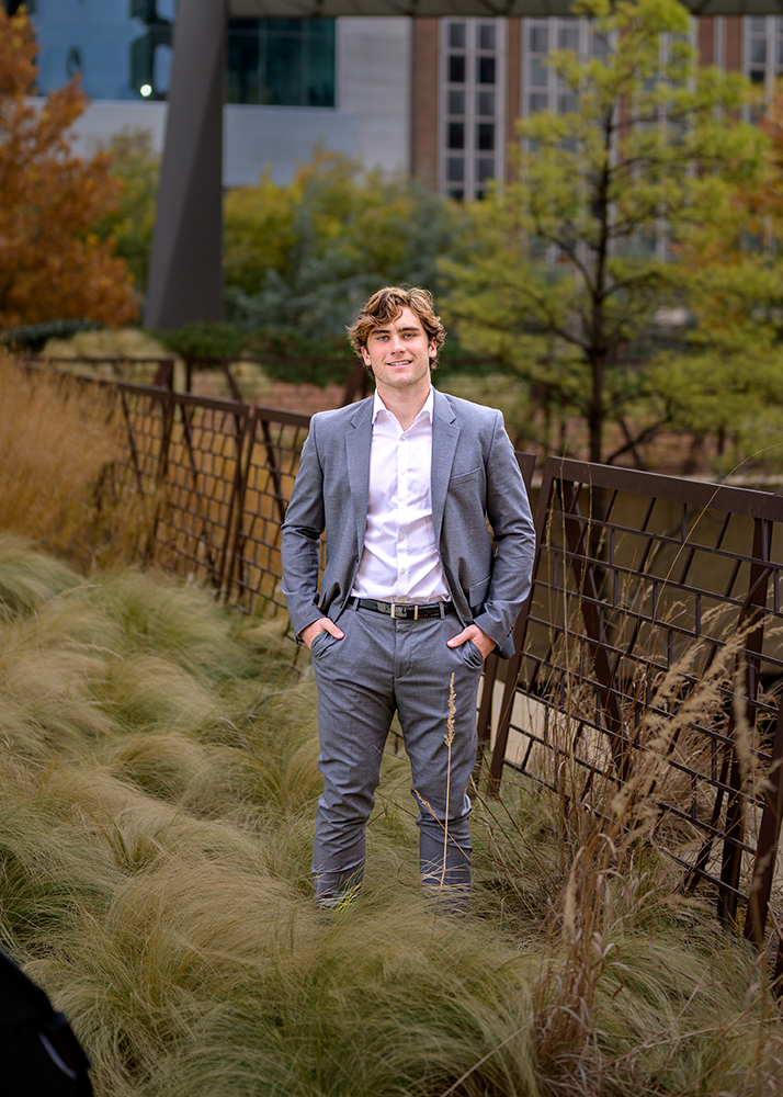A high school senior in a gray suit stands confidently with hands in pockets, surrounded by tall grass in downtown OKC. Trees and modern buildings form the backdrop, conveying a serene yet professional tone.