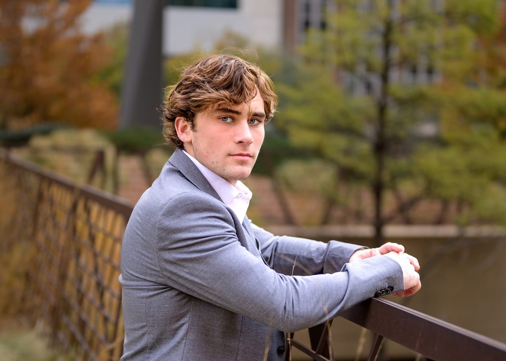 Young man with light brown hair in a gray suit leans on a railing outdoors. The background is blurred with green and brown trees, conveying a calm tone.