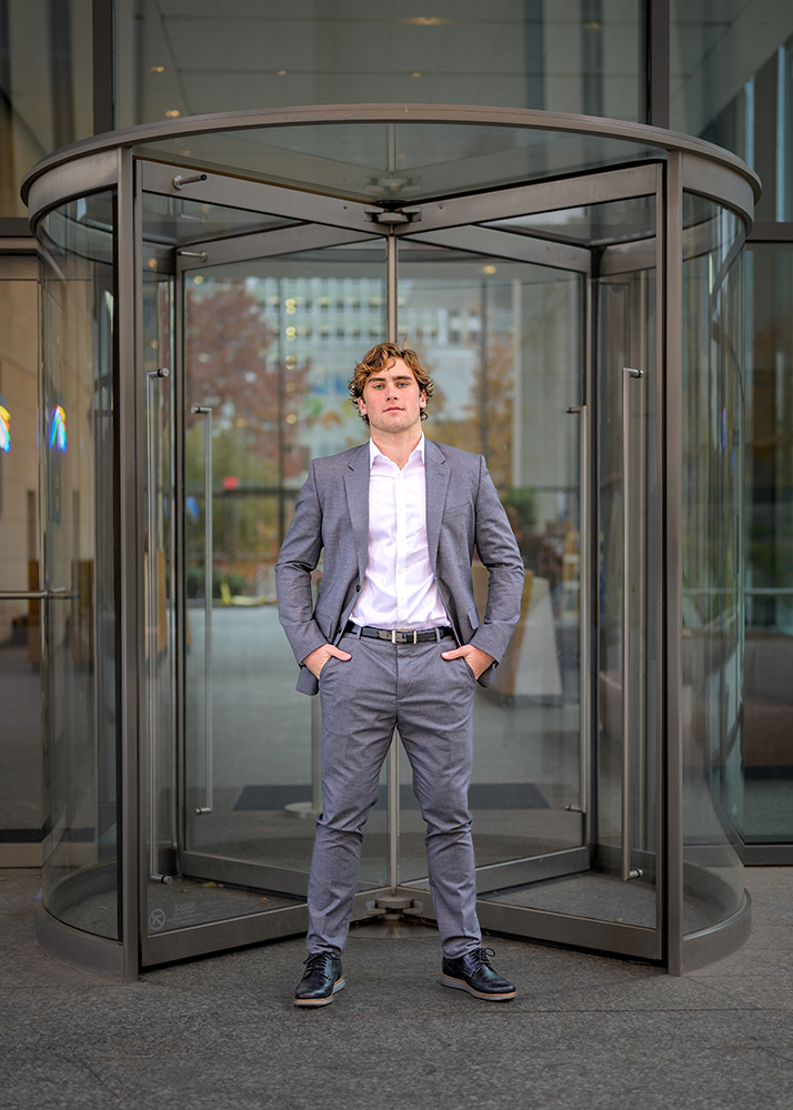 Senior boy in a gray suit and white shirt stands confidently in front of a revolving glass door, arms at his sides. The background shows an urban setting with tall buildings.