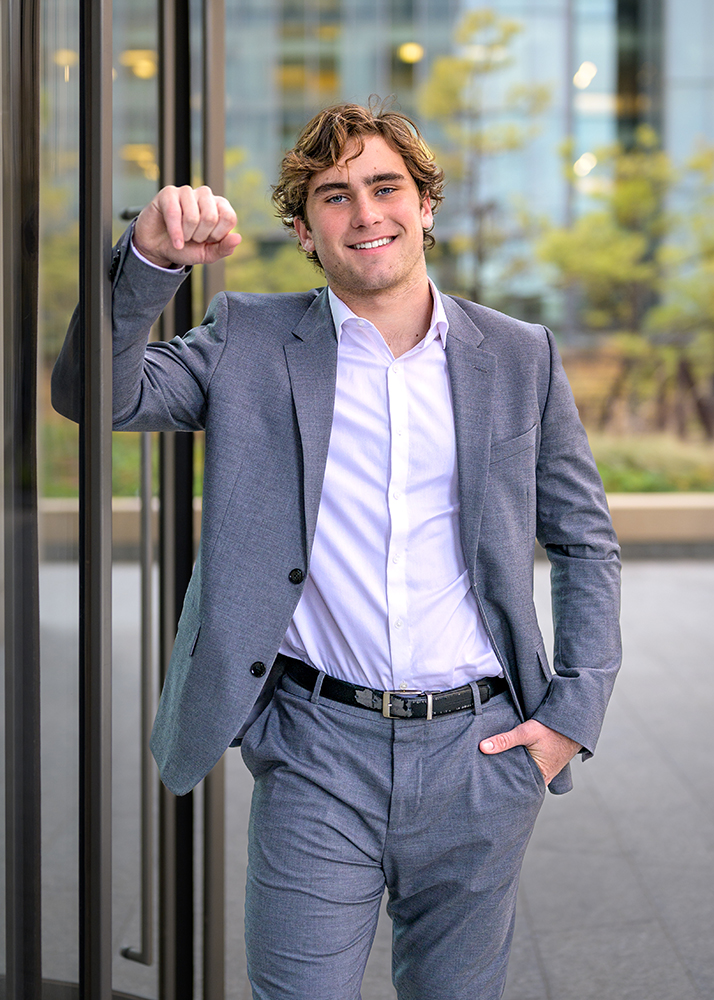 A high school senior in a light gray suit and white shirt smiles confidently while leaning against a glass door outdoors. The background is blurred greenery and modern architecture.