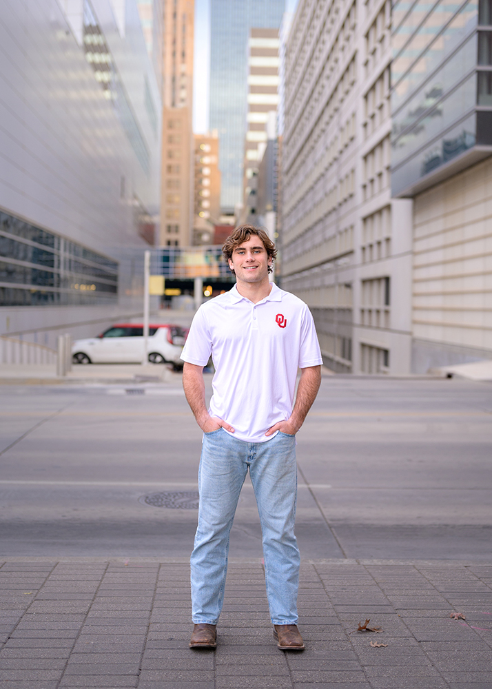 Senior boy in white polo and jeans standing confidently on a city street with tall, modern buildings in the background, conveying urban energy.