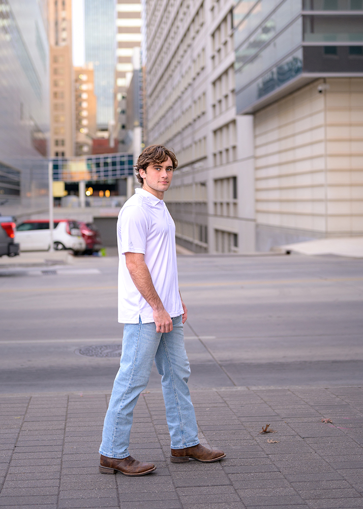 Senior boy in white walking confidently on an OKC street with tall, modern buildings in the background, conveying urban energy.
