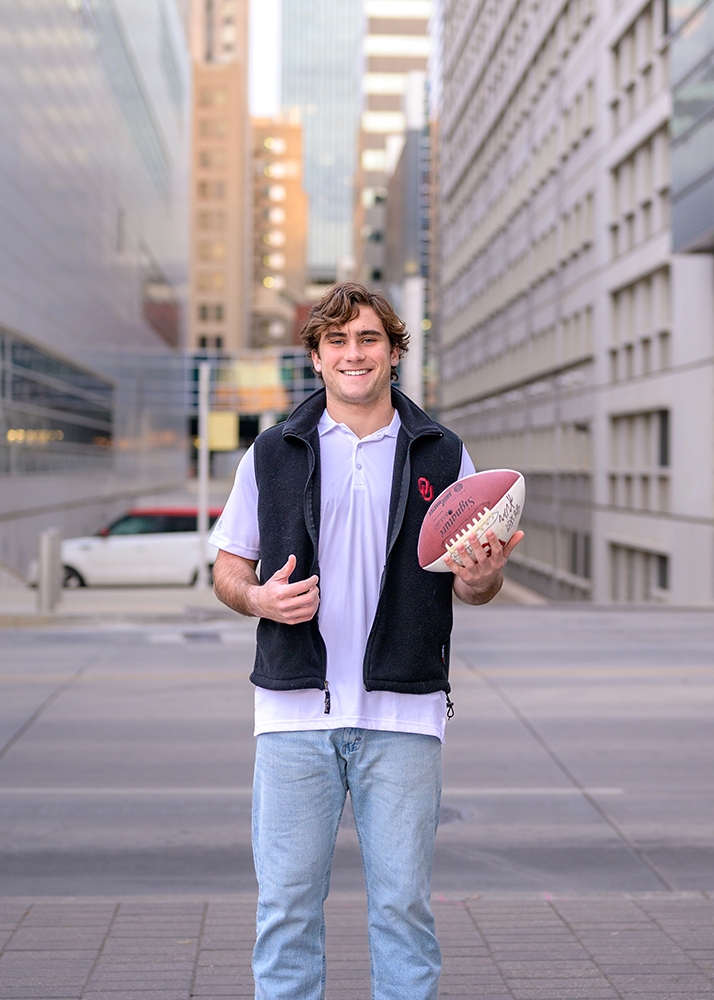 Young man smiling in a city street, wearing a black vest over a white shirt and jeans, holds a football. Background features modern buildings.
