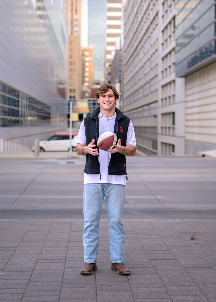Smiling high school senior in casual attire holds a football on a city street flanked by tall buildings. The scene conveys a relaxed urban vibe.