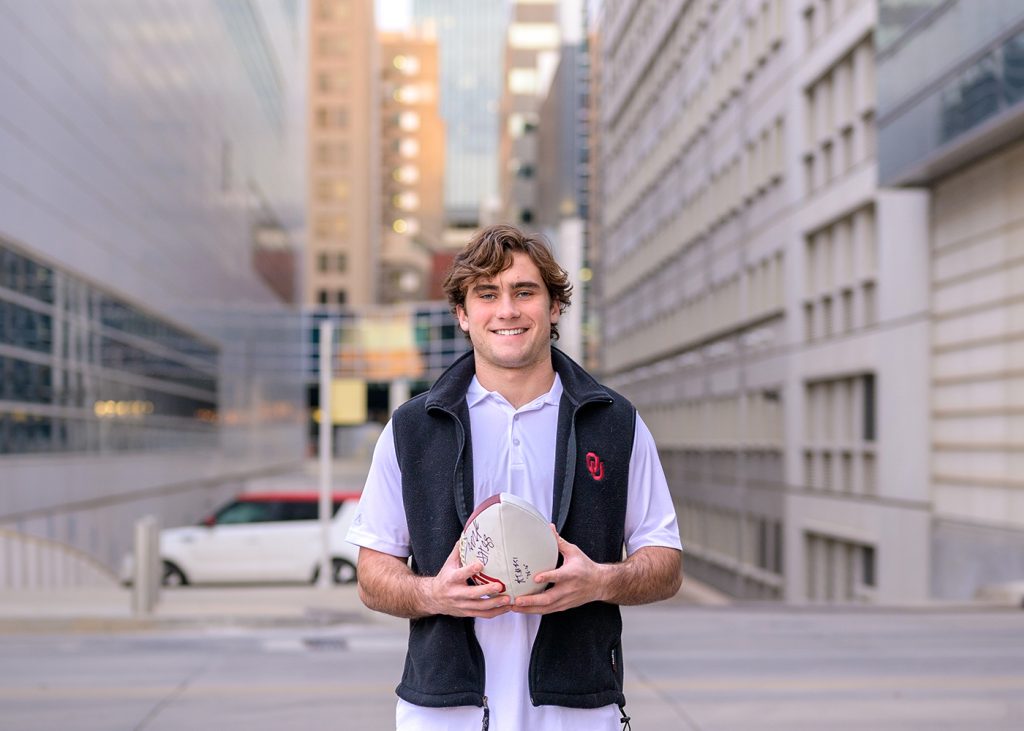 Senior guy smiling on a city street, holding a signed football. Wearing a white polo and black vest with an "OU" logo. Background shows tall buildings.