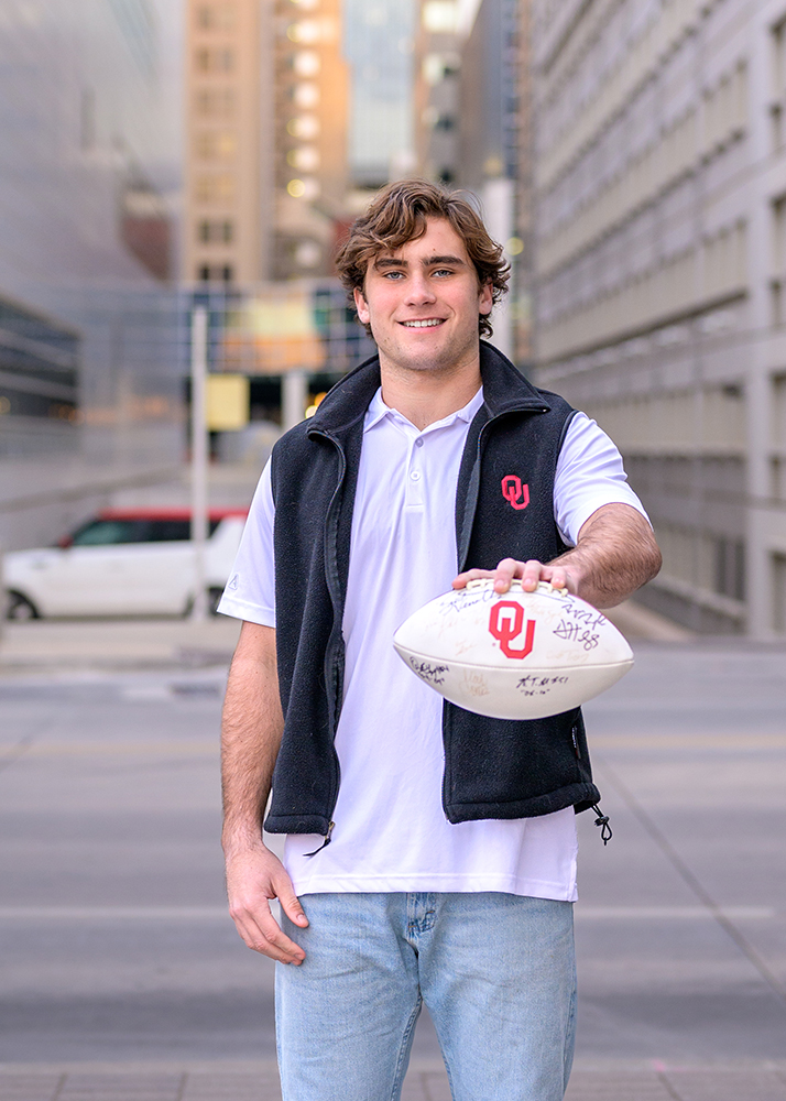 A smiling high school senior stands on a city street, holding a signed football with the University of Oklahoma logo. They're wearing a white shirt and black vest, creating a relaxed and positive atmosphere. Urban buildings blur into the background.