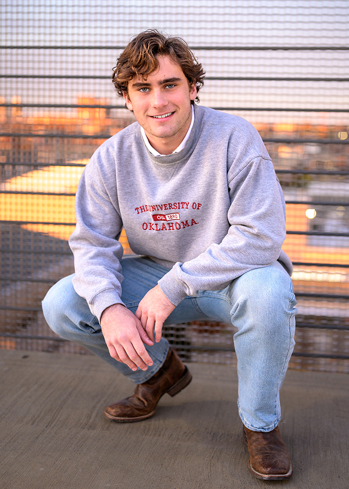High school senior with curly hair crouches, smiling in a gray "University of Oklahoma" sweatshirt and jeans. Background shows an urban rooftop setting.