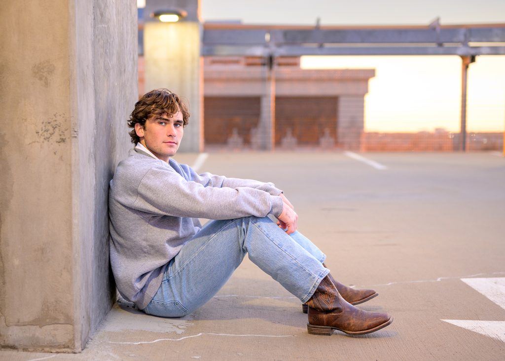 A senior boy wearing a gray sweatshirt, jeans, and brown boots sits against a concrete wall on a rooftop parking lot, looking thoughtfully at the camera.