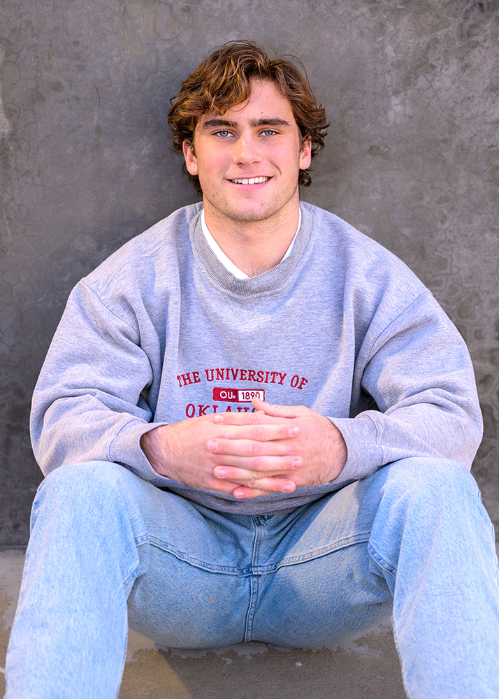 Young man sitting against a gray textured wall, wearing a light gray University of Oklahoma sweatshirt and jeans, smiling confidently. Casual and relaxed mood.