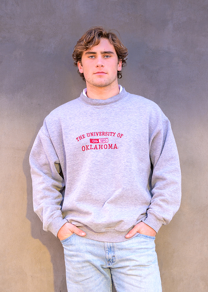 Senior boy in gray "University of Oklahoma" sweatshirt stands against a gray wall, hands in pockets, with a confident and relaxed expression.