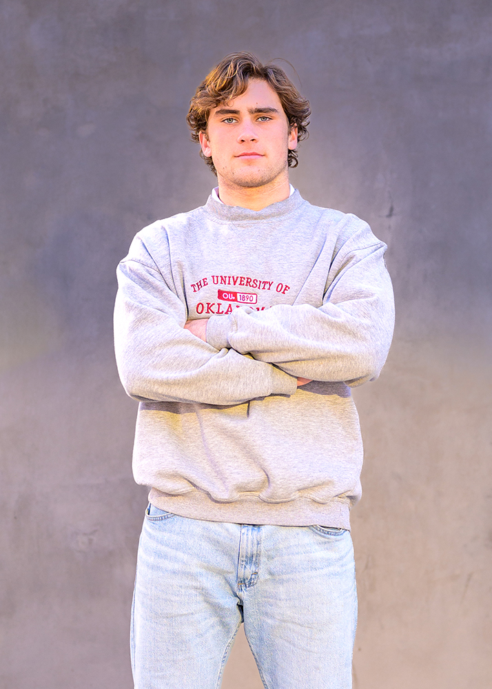 Senior boy in gray "University of Oklahoma" sweatshirt stands against a gray wall, crossing arms, with a confident and relaxed expression.