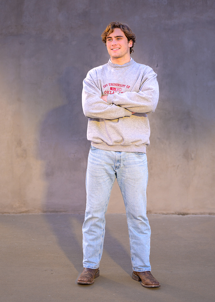 High school senior in gray "University of Oklahoma" sweatshirt stands against a gray wall, crossing arms, with a confident and relaxed expression.