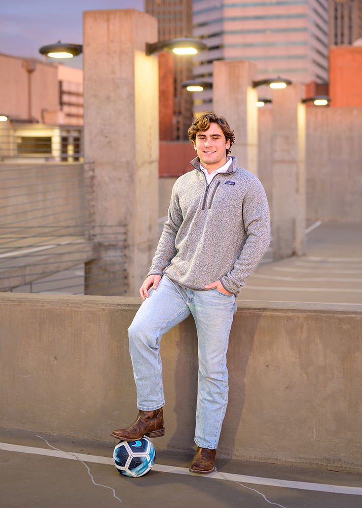 Senior guy in a gray sweater and jeans leans on a low wall in a rooftop parking lot at dusk, smiling and resting a foot on a soccer ball. Oklahoma City background.