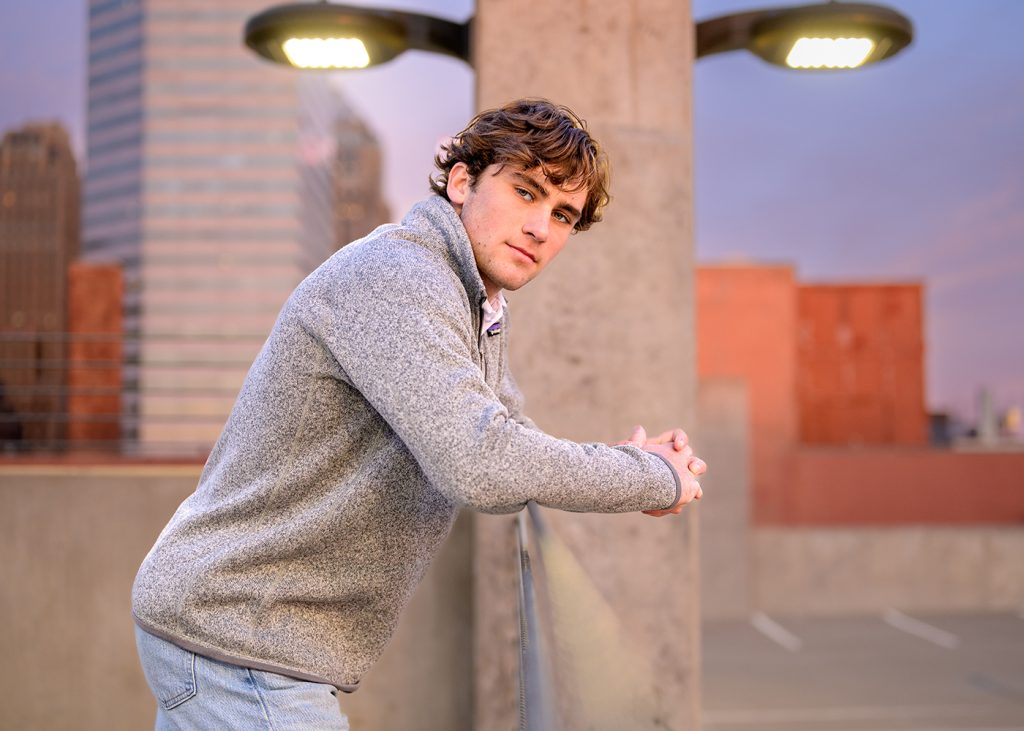 Young man in a gray sweater leans casually on a rooftop railing, city skyline at dusk in the background, exuding a contemplative mood.