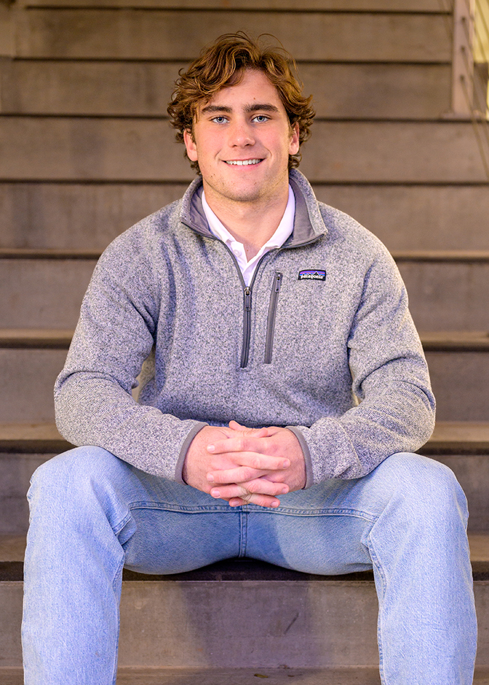 Senior boy with curly hair sitting on steps, wearing a gray fleece and blue jeans, looking at the camera with a relaxed smile and hands clasped.