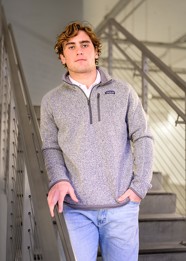 High school senior in a gray zip-up sweater and jeans stands on stairs, hand on railing, with a thoughtful expression. Industrial background.