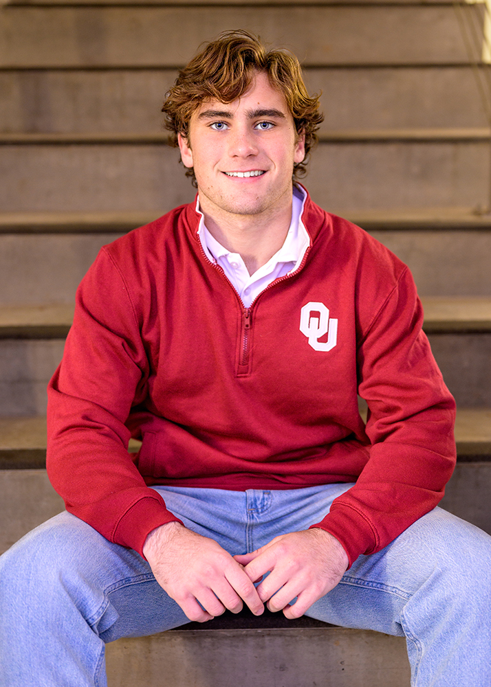 High school senior with curly brown hair sits on stairs, wearing a red OU jacket and jeans. He smiles warmly, conveying a friendly and approachable demeanor.