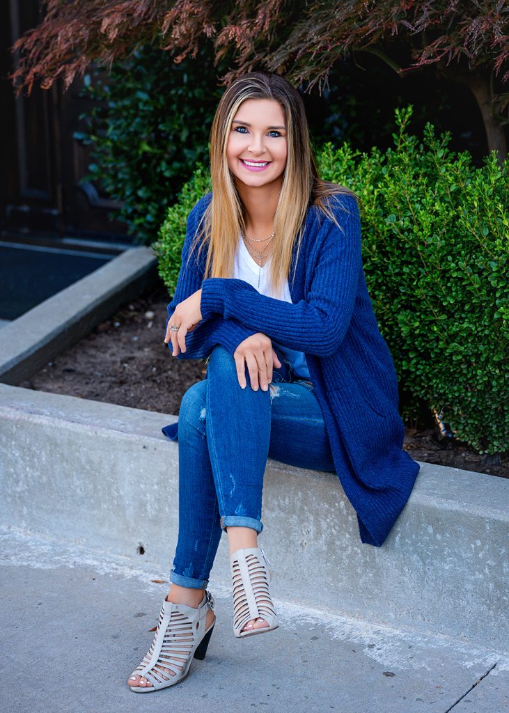 Smiling woman sits on a ledge, wearing a blue sweater, ripped jeans, and white strappy heels