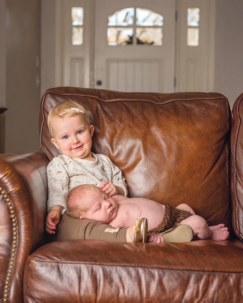 A toddler with a small bow in her hair smiles warmly while sitting on a brown leather couch, gently holding a peacefully sleeping baby on her lap.
