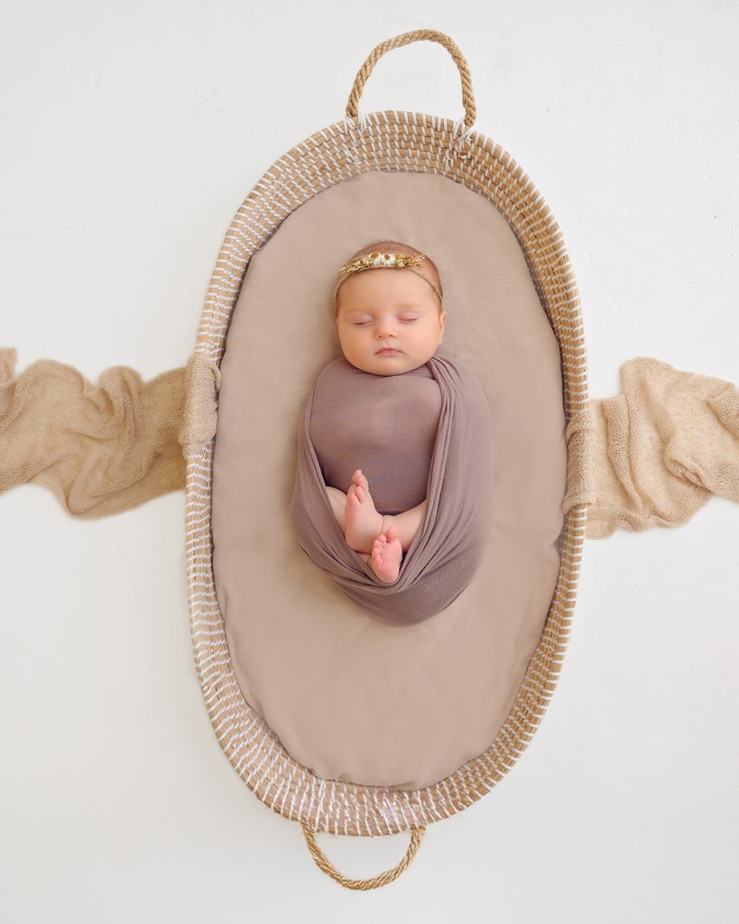 A toddler with a small bow in her hair smiles warmly while sitting on a brown leather couch, gently holding a peacefully sleeping baby on her lap.