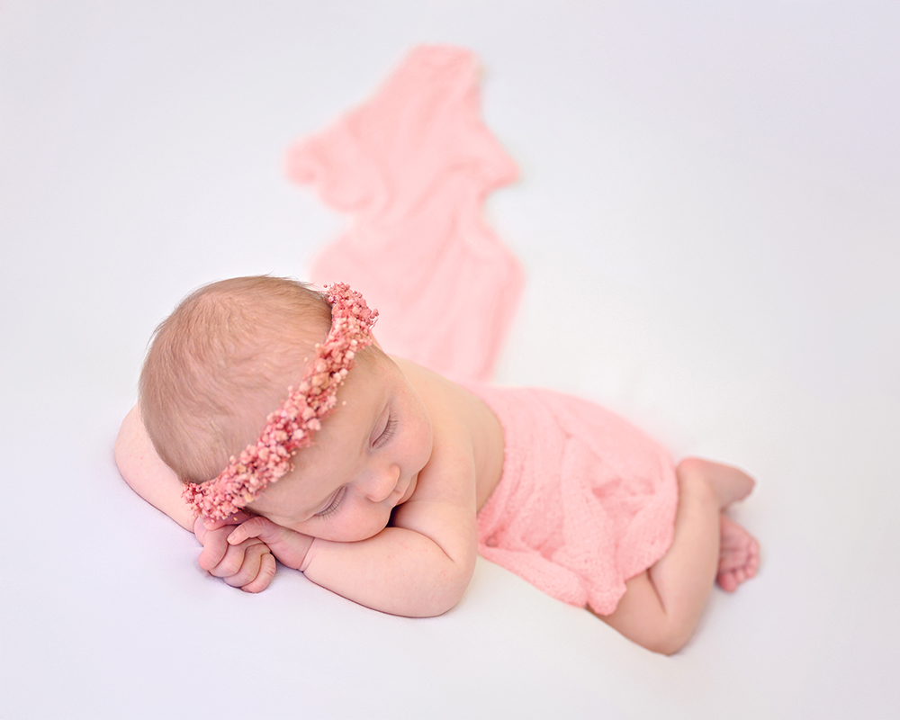 Sleeping baby with a pink flower headband and wrap on a white background, conveying calmness and innocence. Soft pink fabric drapes in the background.