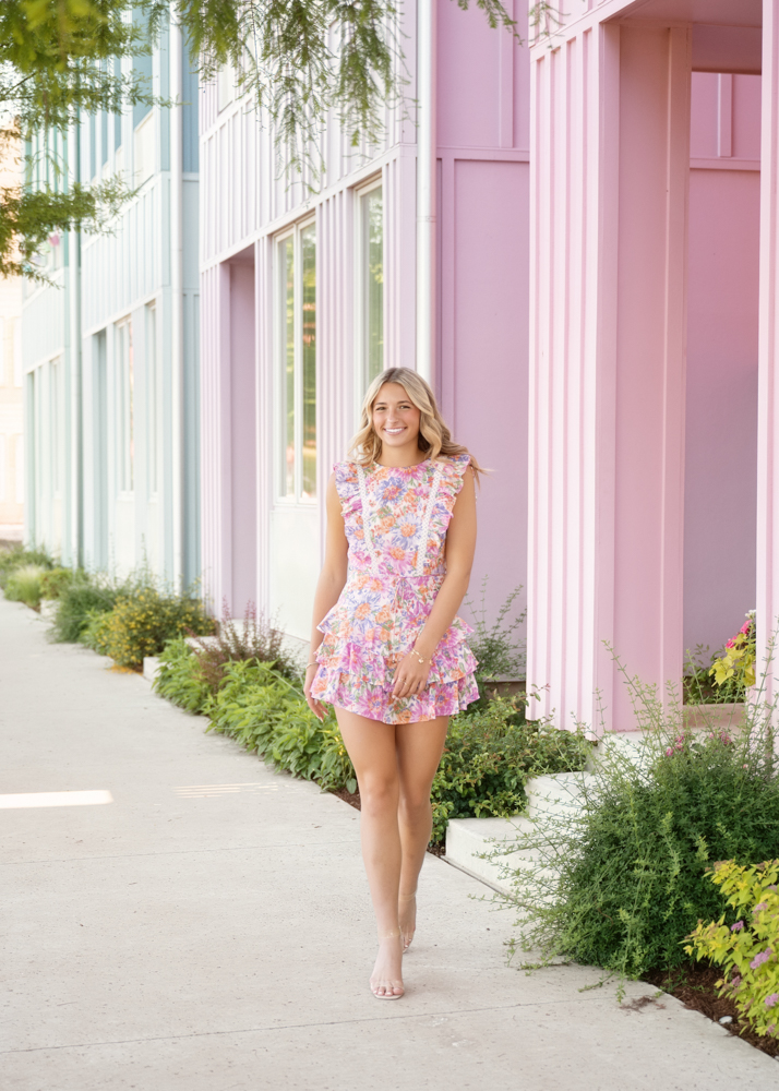 Young woman in a colorful floral dress walks along a sidewalk next to pastel pink and blue buildings, surrounded by lush green plants. She smiles, conveying a cheerful and relaxed atmosphere.