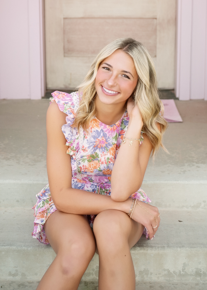 Young woman with long blonde hair sits on steps, smiling warmly. She's wearing a floral dress with ruffles, exuding a cheerful and relaxed vibe.