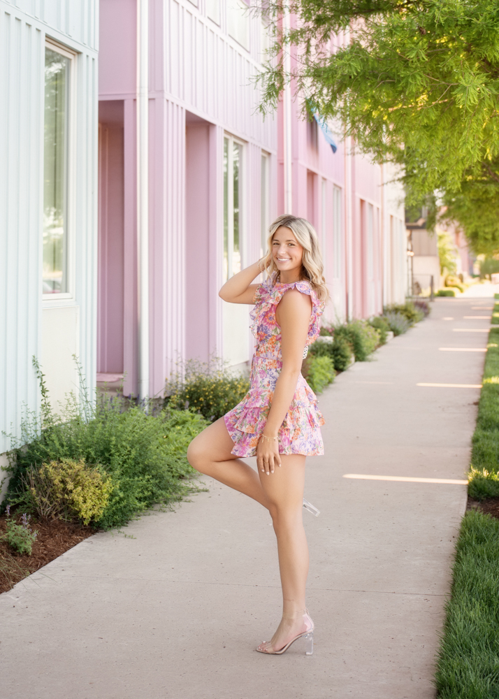 A smiling senior girl poses in a floral dress and heels, standing on a sidewalk beside pastel-colored buildings and lush greenery, exuding joy.