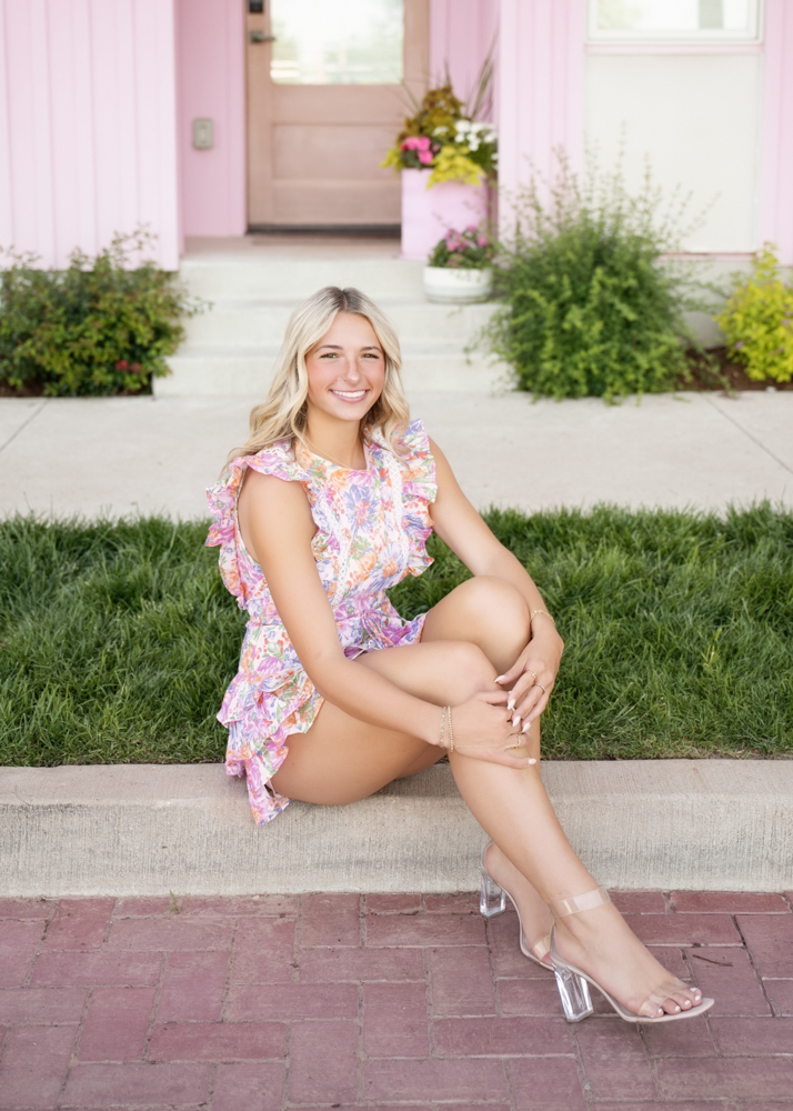 A young woman sits on a curb, smiling warmly. She's wearing a floral ruffled dress and clear heels. Behind her is a pink house with green plants.