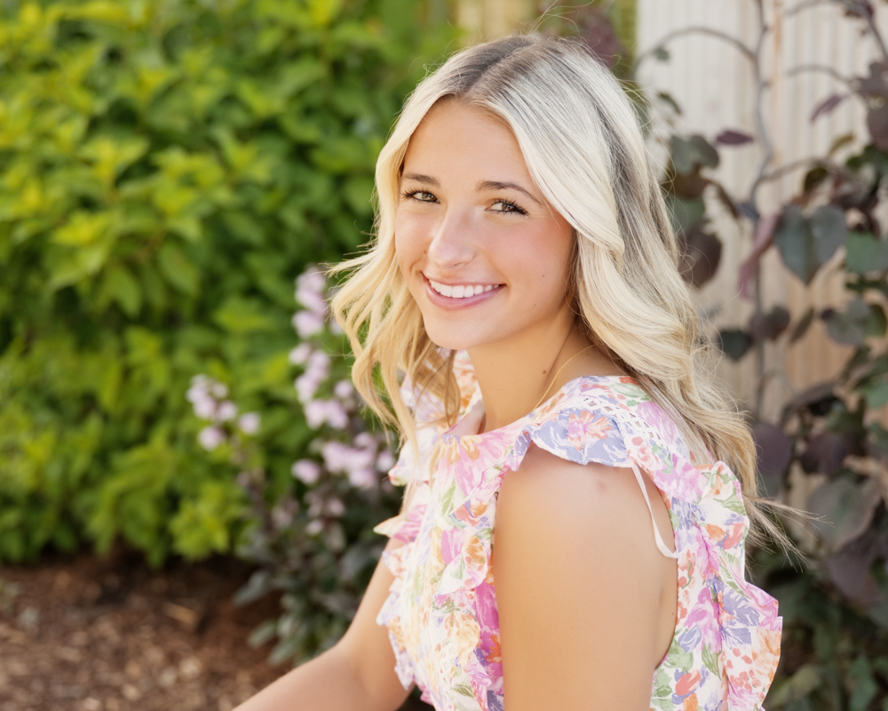 High school senior with long, wavy blonde hair sits outdoors, wearing a floral top. Greenery and purple flowers create a vibrant, cheerful background.