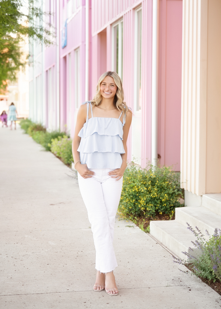 A young woman with long blonde hair smiles, standing on a sidewalk beside pink buildings. She's wearing a light blue ruffled top and white pants, conveying a cheerful and summery vibe.