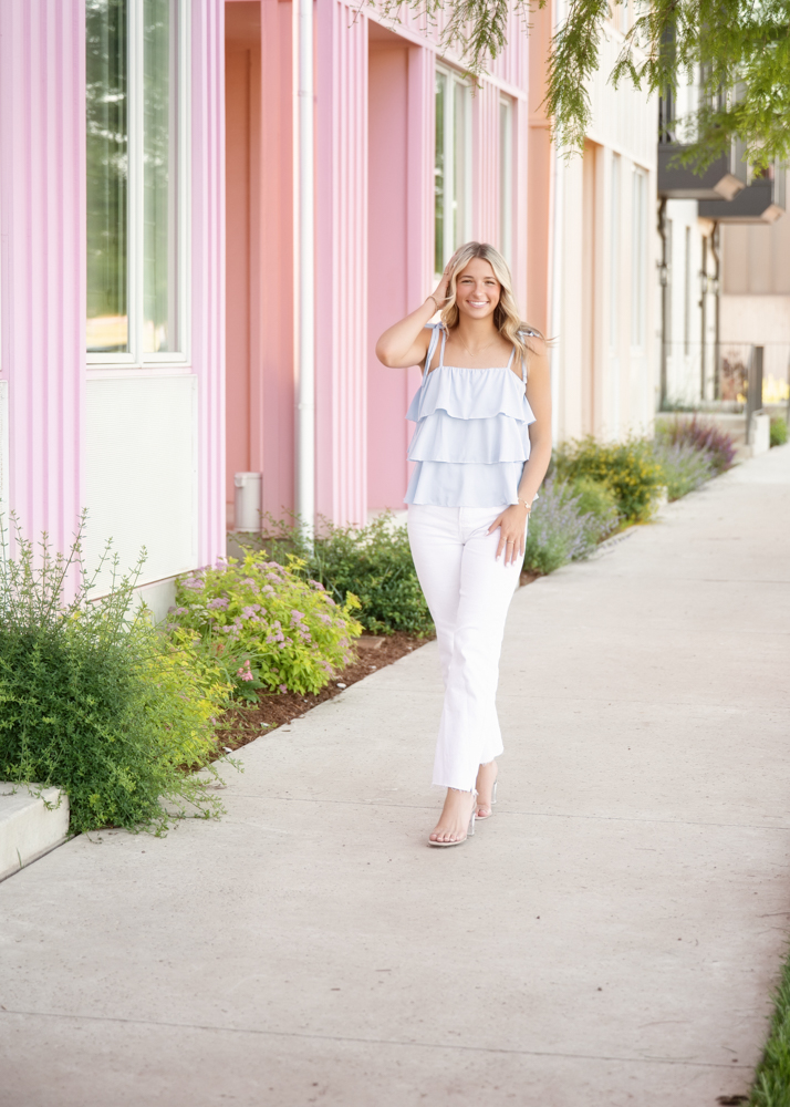 A woman walks along a sidewalk beside pastel pink and white striped walls, smiling. She wears a light blue ruffled top and white pants, exuding a cheerful, casual vibe. Lush greenery borders the path.