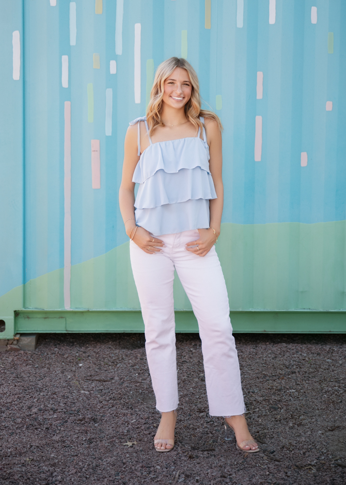 A smiling person stands in front of a colorful, striped wall. They wear a light blue ruffled top and white pants. The scene is bright and cheerful.v