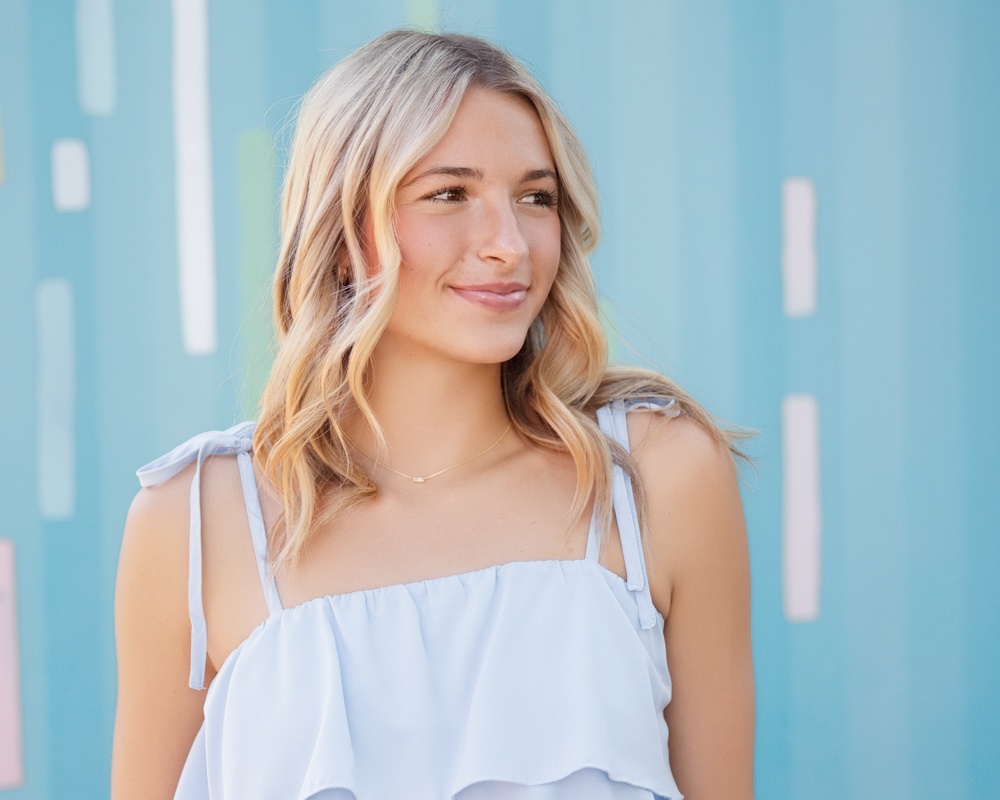A smiling woman with blonde hair stands against a pastel blue wall with colorful vertical stripes. She's wearing a light blue dress, conveying a cheerful and relaxed mood.