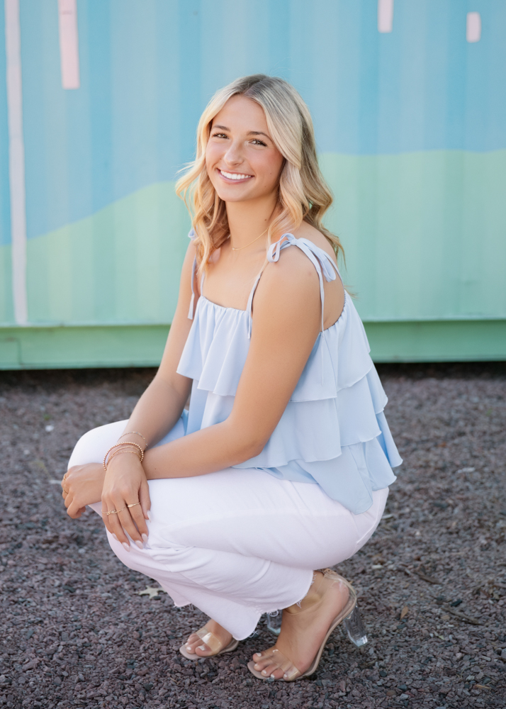 A smiling woman crouches on gravel, wearing a light blue ruffled top and white pants. The background features a pastel blue and green wall, conveying a cheerful atmosphere.