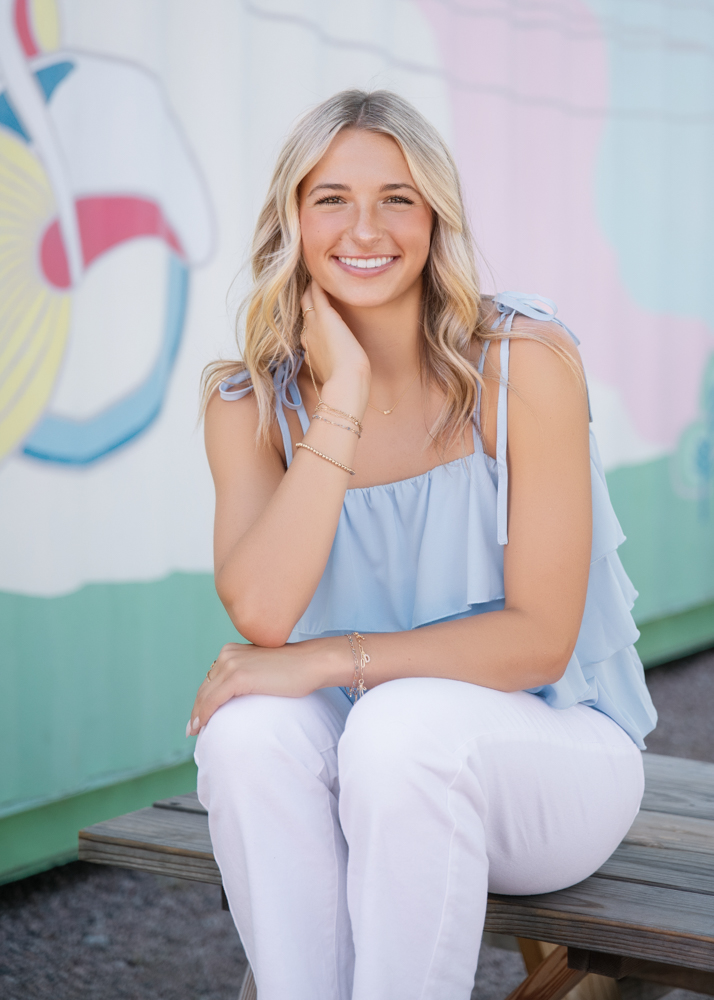 A smiling woman in a light blue ruffled top and white pants sits on a wooden bench. Colorful mural in the background adds a cheerful vibe.