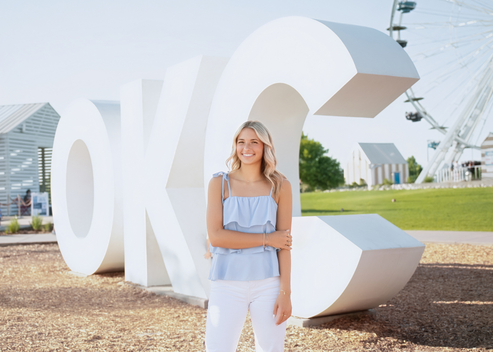 Kenna's senior photo session at Wheeler Park in front of large white "OKC" letters with a Ferris wheel and greenery in the background.