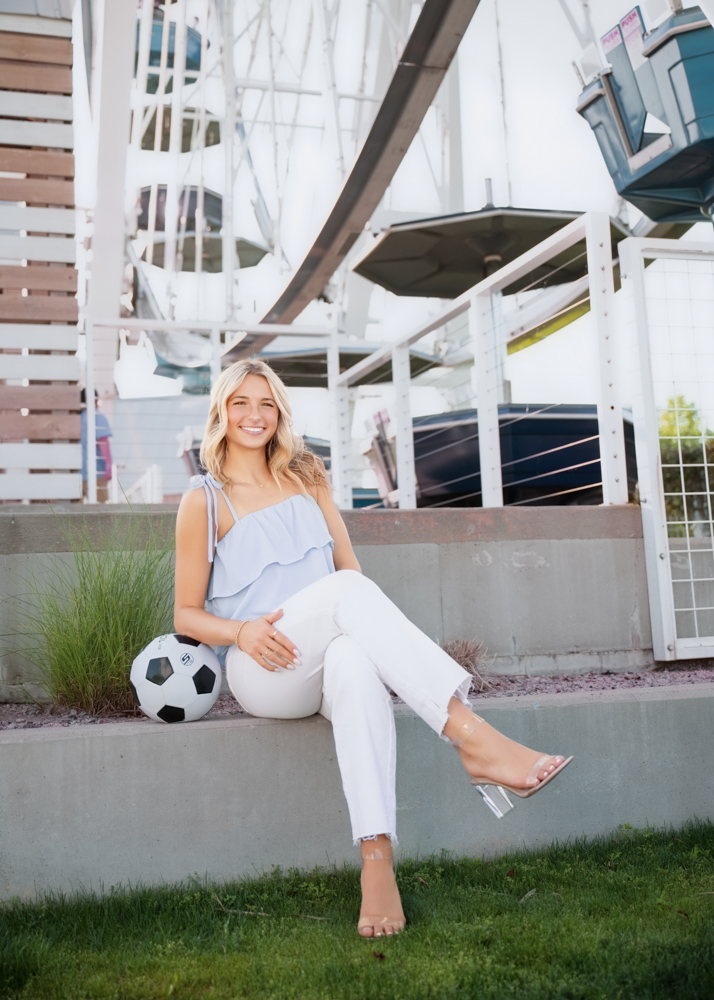 A smiling woman in a blue top and white pants sits on a ledge by a soccer ball. Behind her is a Ferris wheel. The scene is bright and cheerful.