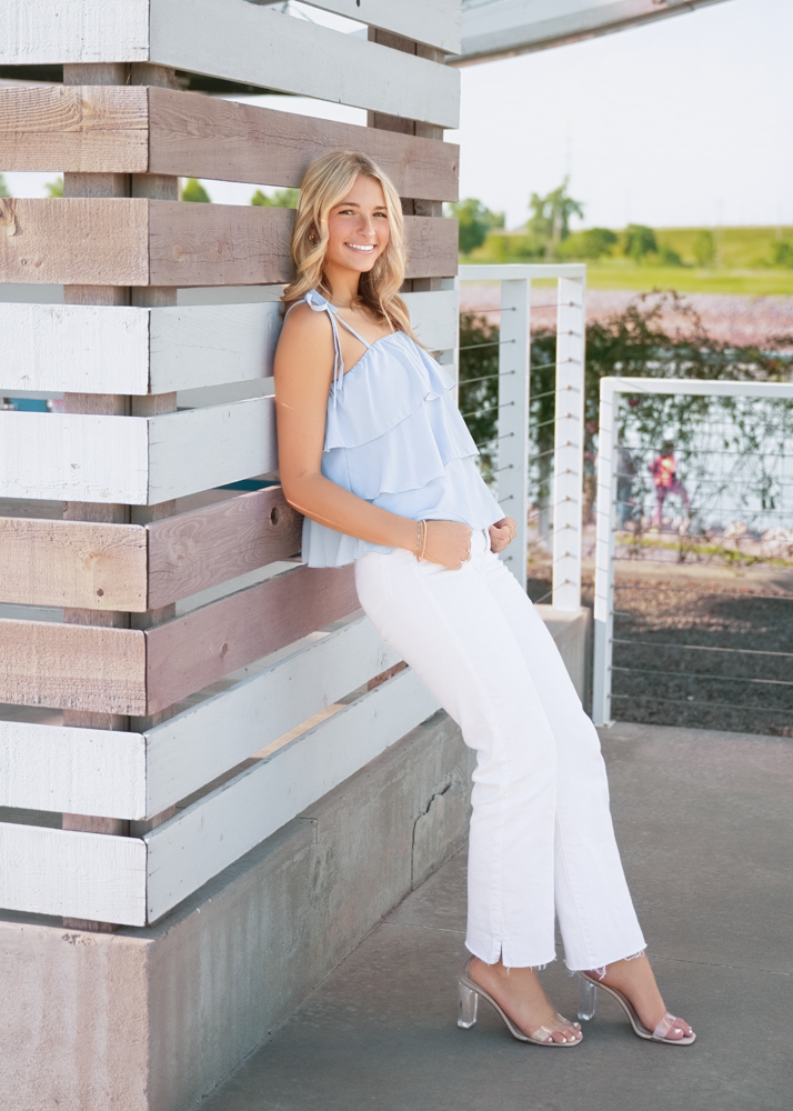 Smiling woman leans against a wooden wall, wearing a light blue ruffled top and white pants. She exudes a relaxed, cheerful vibe in a sunny setting.
