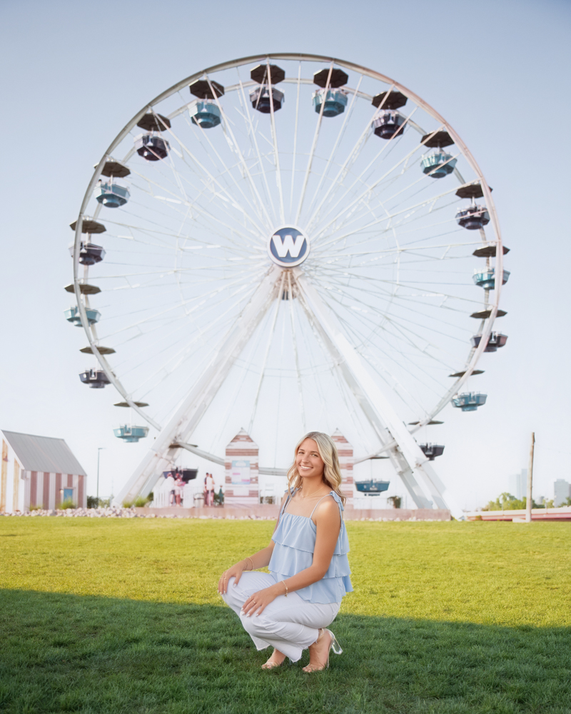 High school senior girl at Wheeler Park in OKC wearing a light blue top and white pants kneels on grass in front of a large Ferris wheel under a clear sky, evoking a cheerful ambiance.