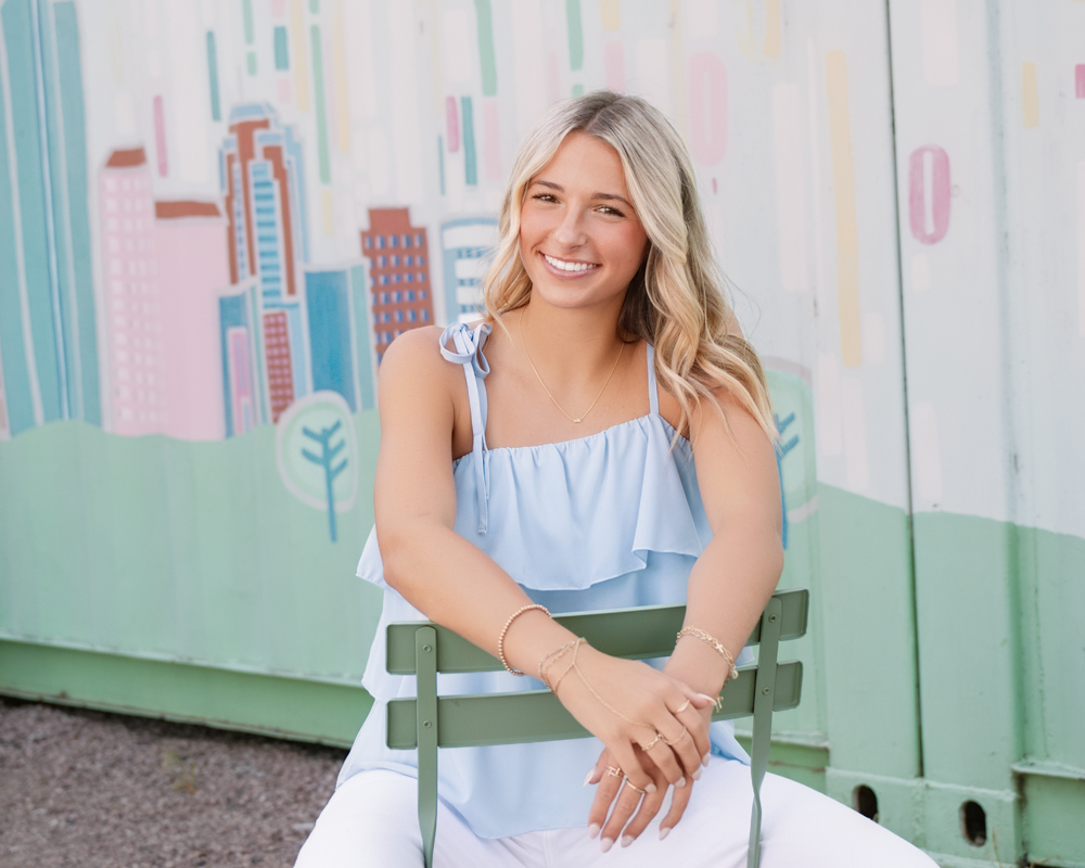 Smiling woman seated on a green chair, wearing a light blue top and white pants. Background features a colorful mural of abstract city buildings.