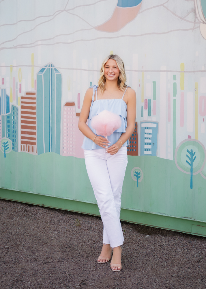 Kenna wears a light blue top and white pants holds pink cotton candy. She stands in front of a colorful cityscape mural, evoking joy.