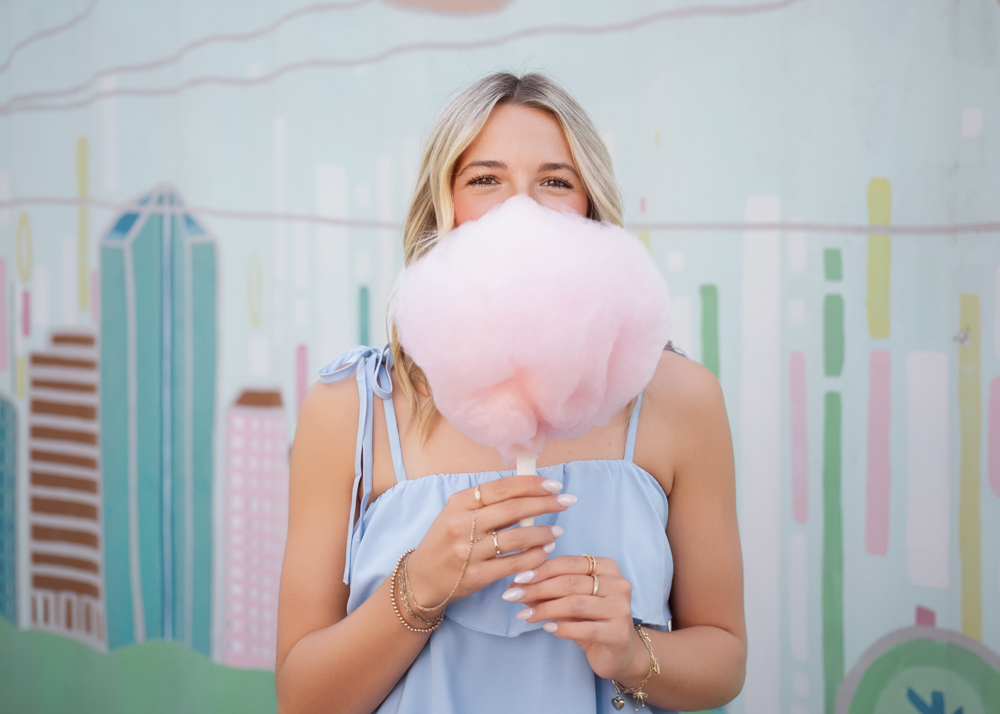 A blonde woman in a light blue top smiles behind a large pink cotton candy, against a pastel urban mural background. Playful and cheerful vibe.